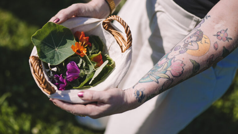 mand met geplukt eetbaar onkruid uit moestuin, vrouw met tattoos op arm houdt mand vast
